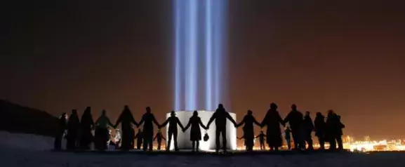 A group of people hold hands around the Imagine Peace tower on Videy island.